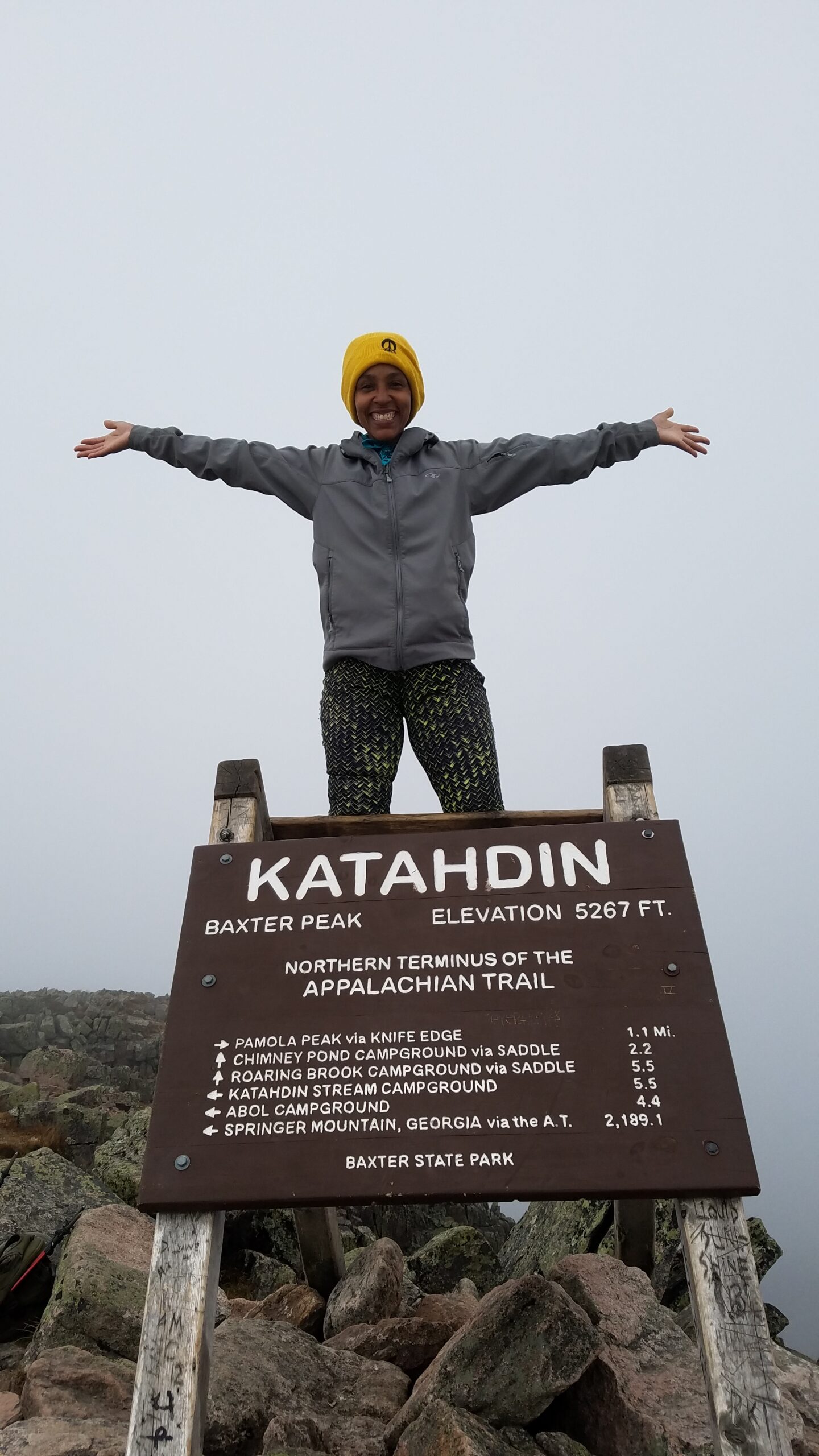 Kimberlee standing with arms outstretched above the Katahdin summit sign at Baxter Peak, the northern terminus of the Appalachian Trail.