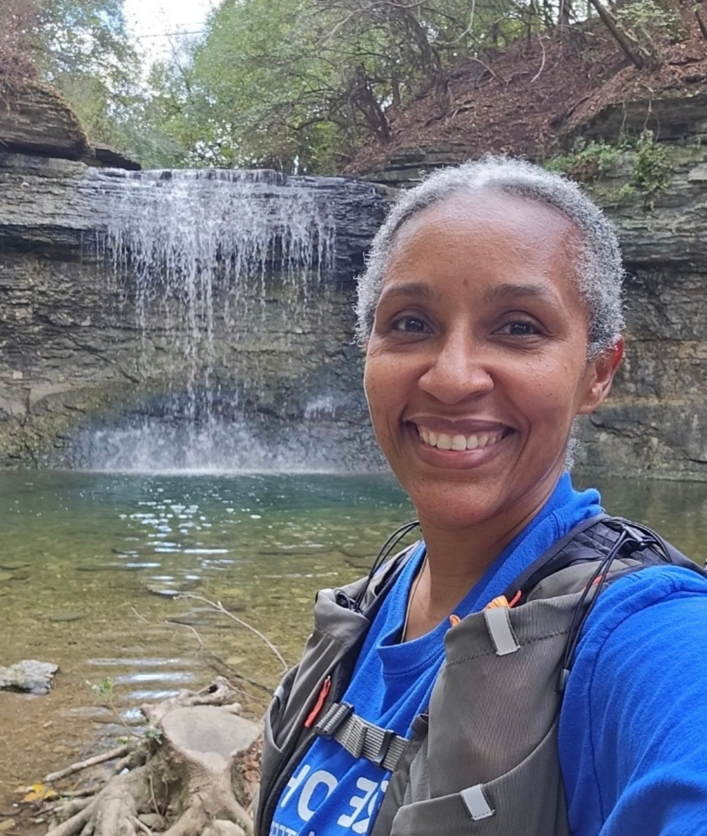Kimberlee smiling beside a waterfall during a hiking adventure, demonstrating her passion for walking, outdoor exploration, and helping others build a sustainable movement practice.
