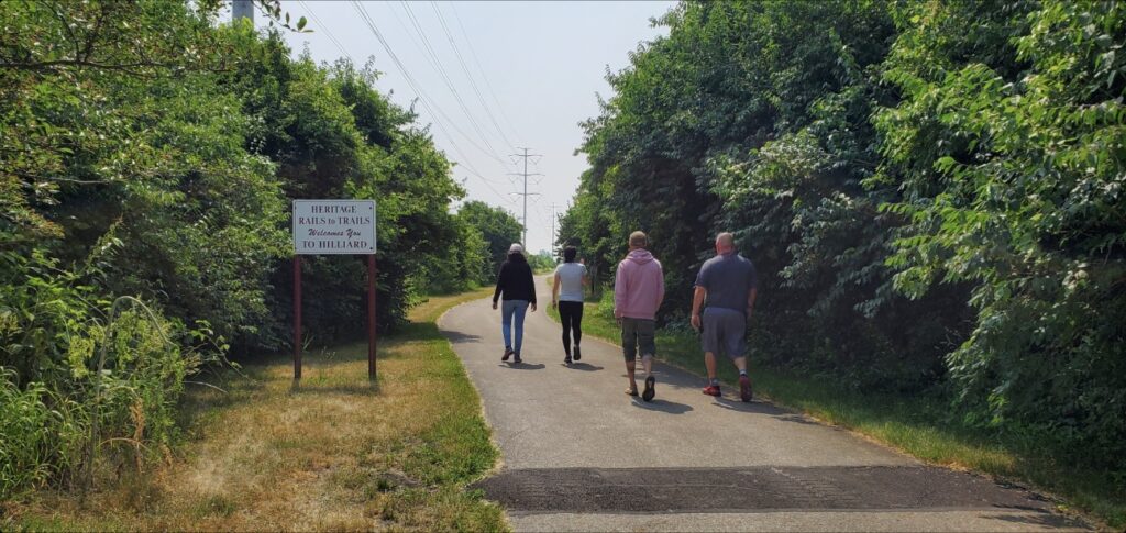 Four people walking along a paved trail surrounded by trees, representing the beginning of a walking wellness journey.